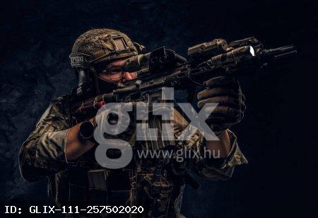 Close-up studio photo against a dark wall. The elite unit, special forces soldier in camouflage uniform holding an assault rifle with a laser sight and aims at the targe