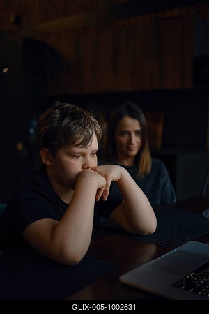 A young boy sits at a table, staring intently at a laptop screen with his hands on his chin. His mother watches him closely from behind, both in a cozy, dimly lit room-stock-foto