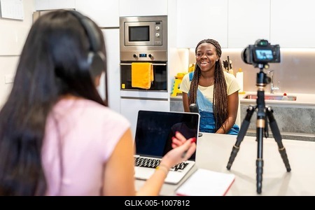 Two teenage girls creating content for their video blog, recording with a camera and using a laptop in a home kitchen-stock-foto