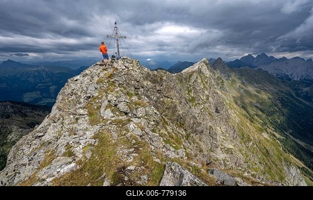 Hiker on the summit of the Raudenspitze with summit cross, narrow rocky ridge, dramatic cloudy sky, Carnic Alps, Carinthia, Austria-stock-foto