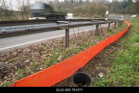 Common toads (Bufo Bufo) in a bucket buried in the ground next to an amphibian fence in the colour orange, protective fence, barrier next to a busy road with crash barrier and delineator, exit of an motorway, moving vehicles, transporter, truck, truck passes by, motion blur, wipe effect, protection, rescue, amphibian migration, toad fence, toad migration, species protection, animal welfare, behaviour, danger, caught, trap, Lower Saxony, Germany-stock-foto