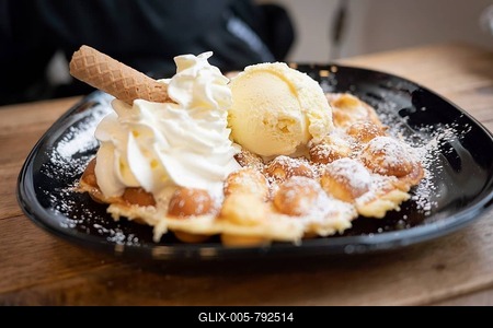 Belgian waffle with vanilla ice cream and whipped cream, waffle rolls, arranged on a black plate and dusted with icing sugar, on a wooden table, in the background a person dressed in black is recognisable, oblique frontal view, Wallonia, Belgium-stock-foto