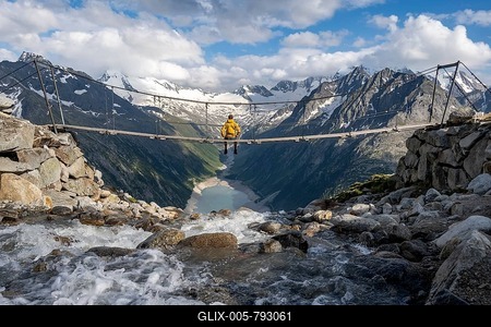 Mountaineer sitting on a suspension bridge over a mountain stream Alelebach, picturesque mountain landscape near the Olpererhütte, view of turquoise blue lake Schlegeisspeicher, glaciated rocky mountain peaks Hoher Weißzint and Hochfeiler with glacier Schlegeiskees, Berliner Höhenweg, Zillertal Alps, Tyrol, Austria-stock-foto