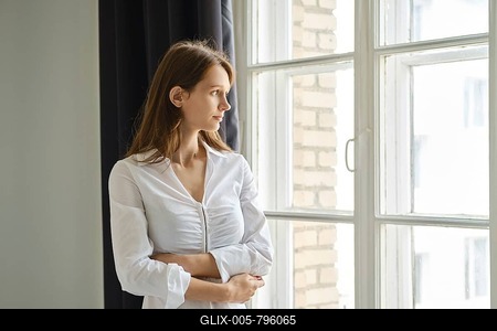 A teenager stands by a window, gazing outside with a thoughtful expression. The cozy room features light curtains and a brick wall, creating a serene atmosphere for reflection and contemplation-stock-foto