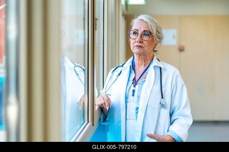 Thoughtful senior female doctor wearing lab coat and stethoscope looking through window in hospital corridor-stock-foto