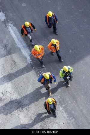 Several construction workers wearing hard hats walk over tarmac. They cast long shadows. Stuttgart, Baden-Württemberg, Germany-stock-foto