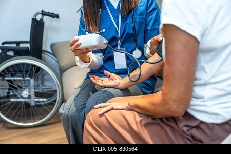 Healthcare professional checking a person's blood pressure with a cuff and digital monitor, providing medical consultation during a home visit with a wheelchair visible-stock-foto