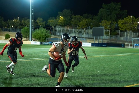 American football player vigorously running with the ball on a green field during a night game, two opposing players chasing in pursuit, highlighting action and competitive athletic performance-stock-foto