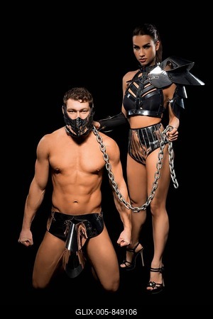 Kneeling muscular male in mask held on chain by dominant brunette female in black BDSM outfit on black background-stock-foto