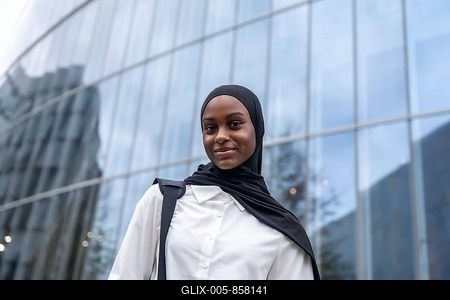 African american woman student wearing a black hijab and white shirt, carrying a backpack, standing confidently in front of a modern glass building in an urban environment-stock-foto