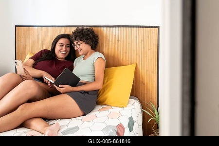 A joyful couple relaxes on a cozy bed. They are sharing a moment, with one reading a notepad and the other using a smartphone, showcasing a casual, happy indoor scene-stock-foto