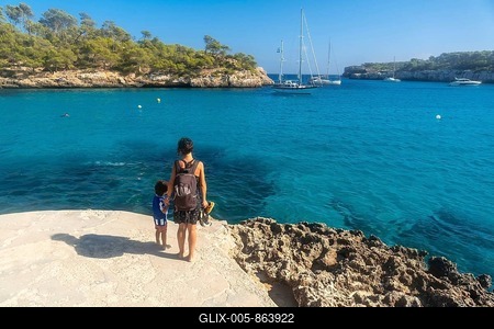 Mother and son standing on a cliff, enjoying breathtaking view of sailboats anchored in the turquoise waters of cala s'amarador, mallorca, spain-stock-foto