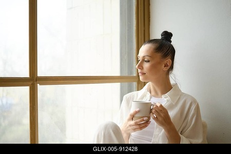 Calm and peaceful woman in white shirt sitting on the windowsill with a cup of tea or coffee in her hands-stock-foto