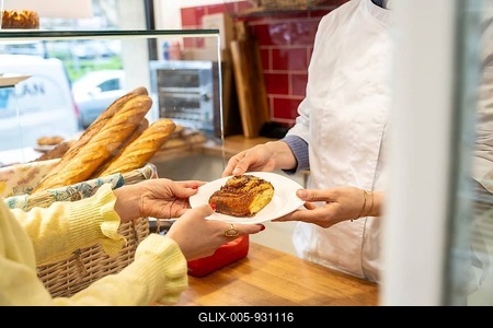 Baker's hands in a white coat serving a freshly baked brioche cinnamon roll on a white plate to a customer at a counter in a busy bakery, highlighting sweet indulgence and excellent service-stock-foto
