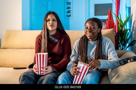 Two teenage girls enjoying a movie night at home, sitting on a comfortable sofa, eating popcorn from striped boxes, and having a great time together-stock-foto