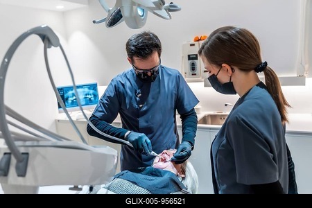 Dentist wearing magnifying glasses and face mask performing a dental procedure on a senior female patient assisted by a dental nurse-stock-foto