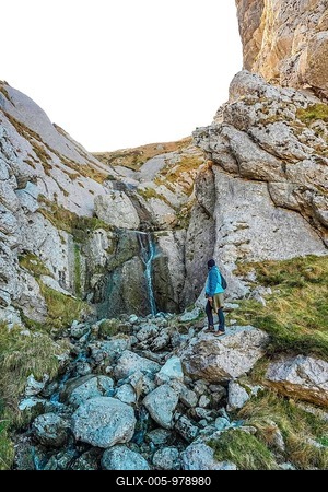Hiker in blue jacket navigating rocky landscape near a small waterfall on mount txindoki, showcasing the rugged beauty of gipuzkoa, basque country-stock-foto