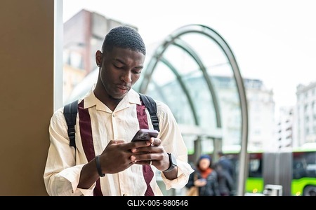 Young african american man with backpack focused on smartphone while waiting at a city bus stop, commuting and browsing apps in a modern urban downtown setting-stock-foto