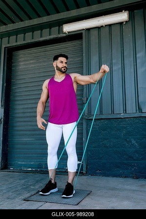 A fit man in a pink tank top and white leggings exercises with a resistance band outdoors. He stands confidently against an industrial background, showcasing fitness and strength-stock-foto