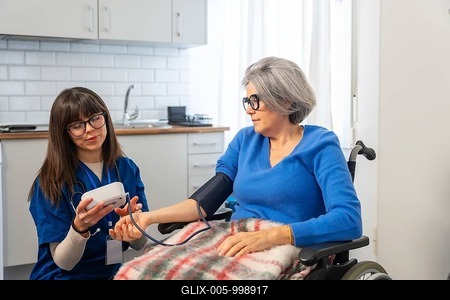 Nurse using a digital device to check blood pressure on arm of an elderly woman seated in a wheelchair, providing healthcare support and monitoring vital signs during a home visit-stock-foto