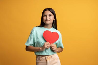 Nettes br?nettes Teenie-M?dchen, das ein rotes Papierherz in den H?nden h?lt und auf gelbem Hintergrund posiert.-stock-photo