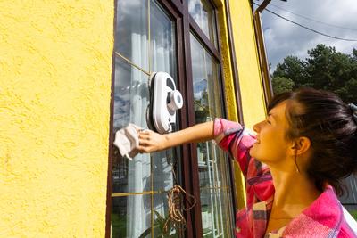 Eine Frau startet einen Fensterputzroboter des Hauses mit gelber Fassade. Haushaltsroboter, Haushaltshilfe, Reinigungsservice-stock-foto