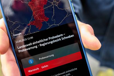 Langweid, Bavaria, Germany - March 13, 2025: Nationwide test alarm in Bavaria. A man holds a smartphone with a warning m-stock-foto
