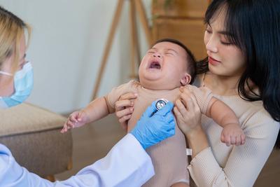 Besorgter Arzt untersucht weinendes Baby im Beisein der Mutter bei einer p?diatrischen Vorsorgeuntersuchung-stock-foto