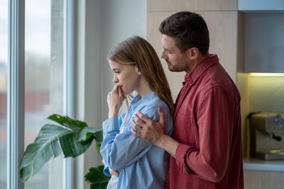 Supportive husband hugging his upset, depressive wife from behind at window in bright apartment Supportive husband huggi-stock-foto