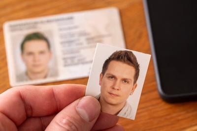 Bavaria, Germany - April 16, 2025: A hand holds a passport photo in front of an ID card and cell phone on a wooden table-stock-foto