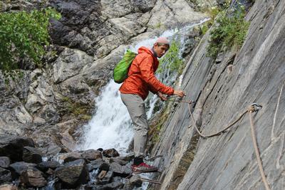 Ein junger Mann in einer leuchtend orangefarbenen Windjacke geht einen schwierigen Bergpfad entlang. Klettersteig im Altai-Reservat-stock-photo