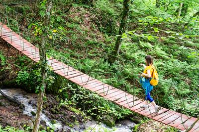 junge Frau mit gelbem Rucksack ?berquert eine schmale H?ngebr?cke ?ber einen rauschenden Bach in einem ?ppigen gr?nen Wald, Konzept des Reisens, der Reisen, des aktiven Lebensstils und der ?berwindung von Hindernissen und Schwierigkeiten im Leben-stock-photo