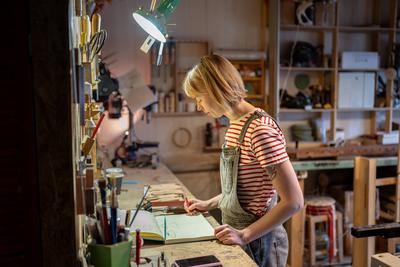 Motivierte Handwerkerin zeichnet Skizzen der geplanten Holzkreationen in ein Notizbuch, bevor sie mit der Arbeit beginnt-stock-photo