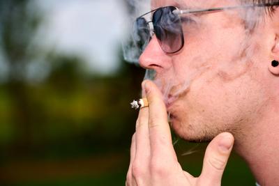 4 May 2024: Man with sunglasses puffs on a cigarette in summer. Smoke from the cigarette spreads over the face *** Mann-stock-foto
