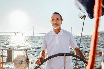 Mature man steering a private yacht while on vacation. Bearded yachtsman standing on his sailing boat looking at distanc-stock-foto