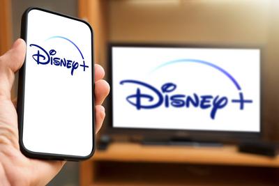 6 April 2024: Man holding a smartphone with Disney Plus logo in front of a TV set with Disney+ video streaming provider-stock-photo