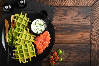 Gr?ne belgische Waffeln. Spinat-, B?rlauch- oder Pesto-Waffeln mit rotem Lachs und Sahnesauce auf schwarzem Teller vor dunklem, rustikalem Holzhintergrund. Leckeres Fr?hst?ck, Snack, Brunch. Ansicht von oben. Attrappe-stock-photo