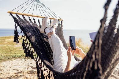 A young woman in casual clothes holds a phone in her hands and lies in a hammock. A girl in a summer hat uses a mobile g-stock-photo