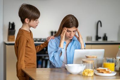 Besorgtes Kind Junge ber?hrt Mutter sitzt am K?chentisch mit Laptop von M?digkeit h?lt Schl?fen-stock-foto