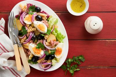 Thunfischsalat mit Nudeln, Eiern, Kartoffeln, Oliven, roten Zwiebeln und Sauce in wei?em Teller auf altem roten rustikalen Tisch Hintergrund. Nicoise Salat. Franz?sische K?che. Ansicht von oben. Flach gelegt.-stock-photo