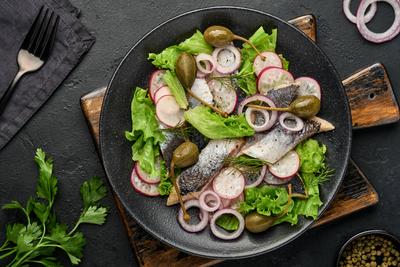 Gesalzener Hering mit Gew?rz, Kapern, Kr?utern und Zwiebelsalat auf schwarzem Teller auf dunklem Hintergrund mit Kopierraum. Marinierter, geschnittener Fisch. Lebensmittel mit gesunden unges?ttigten Fetten, flach gelegt-stock-foto
