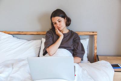 Gedankenreiche junge Frau im Bett mit einem Laptop, ruht ihr Gesicht auf ihrer Hand und sieht besorgt, reflektieren Stress oder ?berarbeit w?hrend Remote Aufgaben Konzept-stock-foto