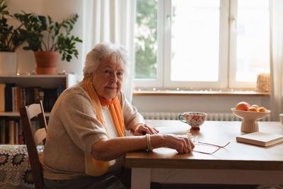 Side view of elderly lady reading a book, sitting at the kitchen table and sipping tea.-stock-foto