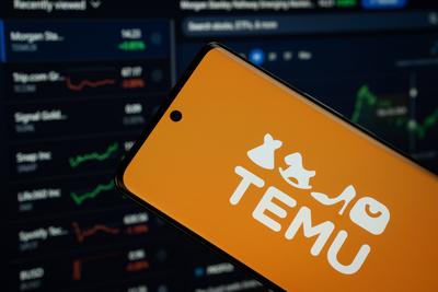 Dhaka, Bangladesch - 18. M?rz 2025: Das Temu-Logo wird auf einem Smartphone-Bildschirm angezeigt, w?hrend im Hintergrund auf einem Computerbildschirm eine Aktiengrafik-Webseite zu sehen ist.-stock-foto