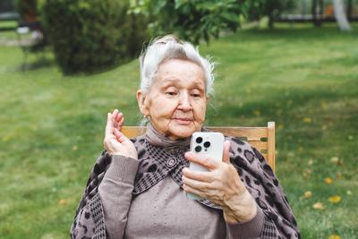 ?ltere Frau sitzt auf einem Stuhl im Freien und winkt w?hrend eines Videogespr?chs ?ber ein Smartphone, Konzept des Alterns und der Technologie-stock-foto