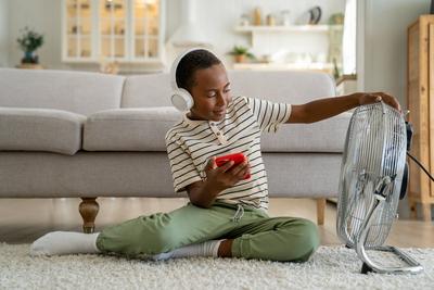 Erfreulich gemischte Ethnie Kind Junge sitzen auf Teppich K?hlung mit Boden elektrischen L?fter h?ren Musik auf Handy-stock-foto
