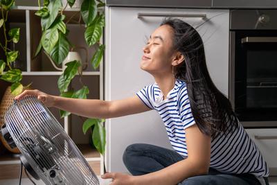 Fr?hliches asiatisches M?dchen genie?t den kalten Wind des elektrischen Ventilators, der in der K?che des Hauses auf dem Boden sitzt-stock-foto