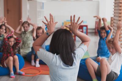 A happy female teacher sitting and playing hand games with a group of little schoolchildren A happy female teacher sitti-stock-foto