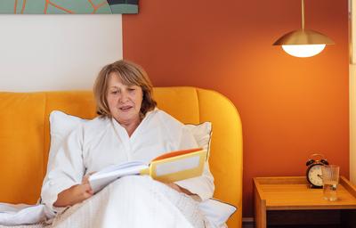 Eine ?ltere Frau in einem wei?en Kittel liest im Bett, bevor sie zu Bett geht. Eine sch?ne Frau mittleren Alters l?chelt und h?lt ein Buch in der Hand. Ein Rentner entspannt sich zu Hause.-stock-foto