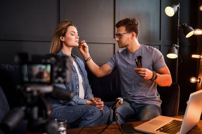 Young male beauty blogger is applying makeup to beautiful young woman Young male beauty blogger sitting on sofa in front-stock-foto